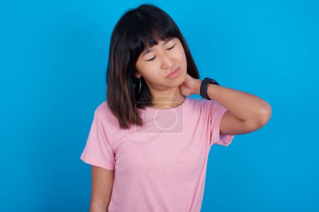 Photo for Young asian woman wearing t-shirt against blue background suffering from back and neck ache injury, touching neck with hand, muscular pain. - Royalty Free Image