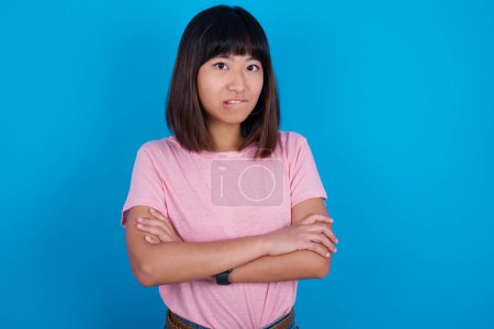 Photo for Young asian woman wearing t-shirt against blue background biting his mouth and looking worried and scared crossing arms, worry and doubt. - Royalty Free Image