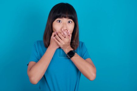 Foto de Aturdido joven asiático mujer usando t-shirt contra azul fondo cubre boca con ambos manos siendo miedo de algo o después de escuchar impresionante chismes. - Imagen libre de derechos