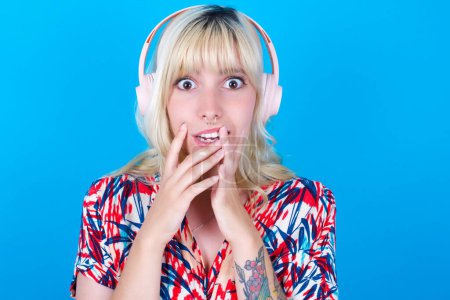 Photo for Shocked caucasian girl wearing floral dress isolated over blue background stares fearful at camera keeps mouth widely opened wears wireless stereo headphones on ears - Royalty Free Image