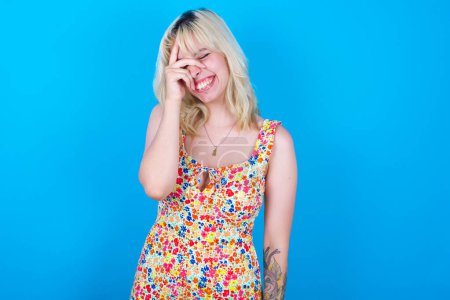Photo for Caucasian girl wearing floral dress isolated over blue background makes face palm and smiles broadly, giggles positively hears funny joke poses - Royalty Free Image