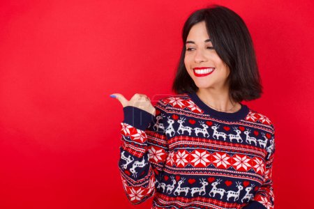 Foto de Encantadora morena mujer caucásica con suéter de Navidad sobre fondo rojo mirando espacio de copia que tiene anuncios - Imagen libre de derechos