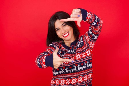 Téléchargez les photos : Brunette femme caucasienne portant chandail de Noël sur fond rouge faisant cadre doigt avec les mains. Concept de créativité et de photographie. - en image libre de droit