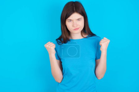 Foto de Chica irritada joven caucásica con camiseta azul aislada sobre fondo azul golpea las mejillas con ira y levanta los puños apretados expresa rabia y emociones agresivas. Modelo furioso - Imagen libre de derechos