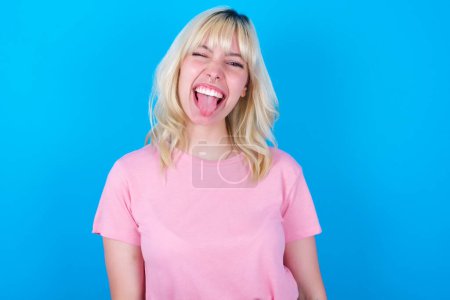 Téléchargez les photos : Fille caucasienne portant t-shirt rose isolé sur fond bleu avec un visage heureux et drôle souriant et montrant la langue. - en image libre de droit