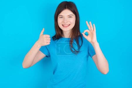 Foto de Joven chica caucásica vistiendo camiseta azul aislada sobre fondo azul sonriendo y luciendo feliz, despreocupada y positiva, haciendo gestos de victoria o paz con una mano - Imagen libre de derechos
