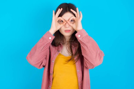 Photo for Playful excited young caucasian girl wearing pink shirt isolated over blue background showing Ok sign with both hands on eyes, pretending to wear spectacles. - Royalty Free Image