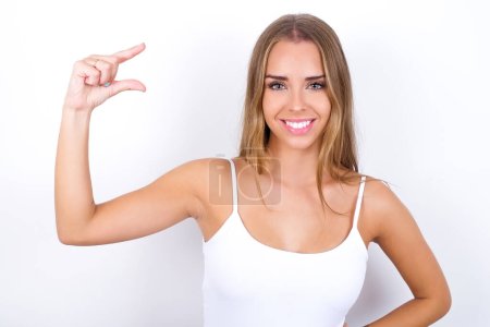 Foto de Joven chica caucásica vistiendo camiseta blanca sobre fondo blanco sonriendo y haciendo gestos con la mano de tamaño pequeño, símbolo de medida. - Imagen libre de derechos