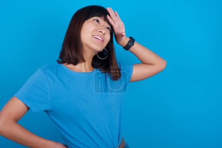 Photo for Young asian woman wearing t-shirt against blue background touching forehead, hears something surprising, glad receive good news, feels relieved. Almost got in trouble. - Royalty Free Image