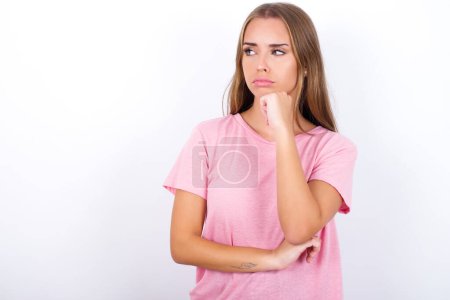 Foto de Joven chica caucásica vistiendo camiseta rosa sobre fondo blanco con la mano bajo la barbilla y mirando de lado con expresión dudosa y escéptica, sospechoso y duda. - Imagen libre de derechos