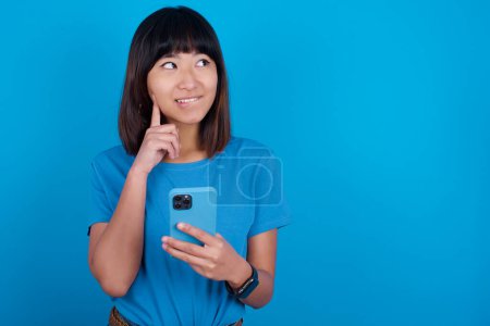 Téléchargez les photos : Image d'une jeune femme asiatique rêveuse portant un t-shirt sur fond bleu à l'aide d'un téléphone portable et tenant la main sur le visage. Prendre des décisions et concept de médias sociaux. - en image libre de droit