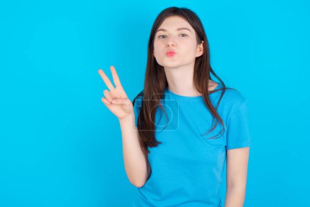 Téléchargez les photos : Jeune fille caucasienne portant T-shirt bleu isolé sur fond bleu fait geste de paix garde les lèvres pliées montre signe v. Concept de langage corporel - en image libre de droit