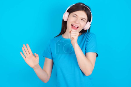 Photo for Happy young caucasian girl wearing blue T-shirt isolated over blue background sings favourite song keeps hand near mouth as if microphone wears wireless headphones, listens music - Royalty Free Image