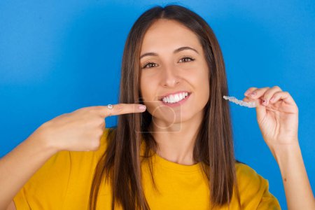 Foto de Hermosa mujer joven sosteniendo un alineador invisible y señalando a sus dientes rectos perfectos. Salud dental y concepto de confianza. - Imagen libre de derechos