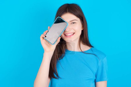 Foto de Joven chica caucásica vistiendo camiseta azul aislada sobre fondo azul sosteniendo teléfono inteligente moderno cubriendo un ojo mientras sonríe - Imagen libre de derechos
