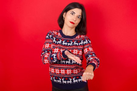 Foto de Morena caucásica mujer usando navidad suéter sobre fondo rojo en prisa apuntando a ver el tiempo, la impaciencia, molesto y enojado por retraso plazo. - Imagen libre de derechos