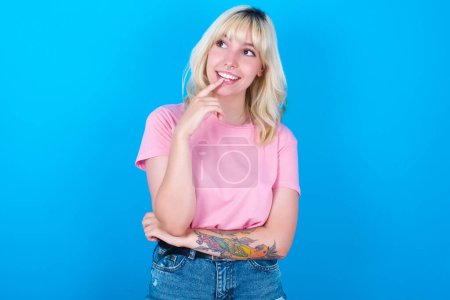 Photo for Caucasian girl wearing pink t-shirt isolated over blue background with thoughtful expression, looks to the camera, keeps hand near face, bitting a finger thinks about something pleasant. - Royalty Free Image