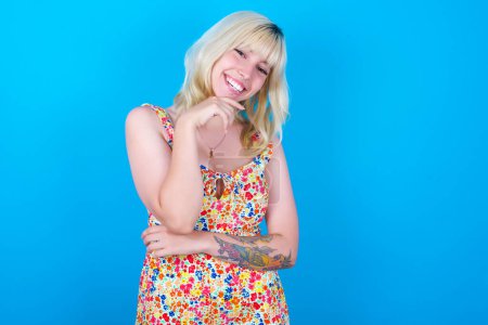 Photo for Caucasian girl wearing floral dress isolated over blue background laughs happily keeps hand on chin expresses positive emotions smiles broadly has carefree expression - Royalty Free Image
