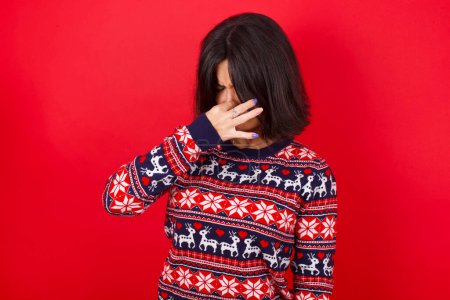 Téléchargez les photos : Brune caucasienne portant un pull de Noël sur fond rouge sentant quelque chose de puant et dégoûtant, odeur intolérable, retenant la respiration avec les doigts sur le nez. Mauvaise odeur - en image libre de droit