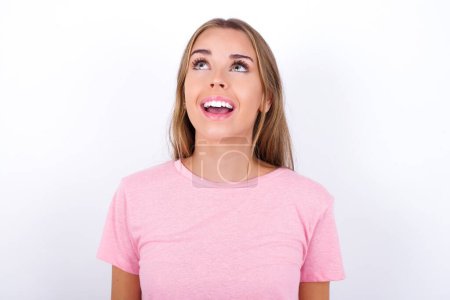 Photo for Surprised Young Caucasian girl wearing pink T-shirt on white background shrugs shoulders, looking sideways, being happy and excited. Sudden reactions concept. - Royalty Free Image