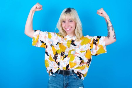 Photo for Waist up shot of caucasian girl wearing floral shirt isolated over blue background raises arms to show muscles feels confident in victory, looks strong and independent, smiles positively at camera. Sport concept. - Royalty Free Image