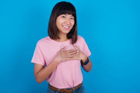 Téléchargez les photos : Honnête jeune femme asiatique portant t-shirt sur fond bleu garde les mains sur la poitrine, touché par compliment ou fait des promesses, regarde la caméra avec grand plaisir. - en image libre de droit