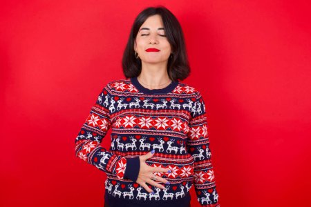 Foto de Morena mujer caucásica vistiendo suéter de navidad sobre fondo rojo toca barriga, sonríe suavemente, comer y concepto de satisfacción. - Imagen libre de derechos