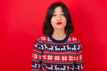 Photo for Shot of pleasant looking brunette caucasian woman wearing christmas sweater over red background pouts lips, looks at camera, Human facial expressions - Royalty Free Image