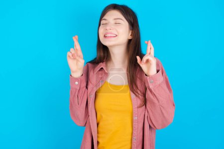 Photo for Joyful young caucasian girl wearing pink shirt isolated over blue background clenches teeth, raises fingers crossed, makes desirable wish, waits for good news, I have to win. - Royalty Free Image