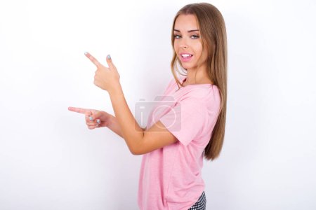 Young Caucasian girl wearing pink T-shirt on white background points aside with  surprised expression with mouth opened, shows something amazing. Advertisement concept.