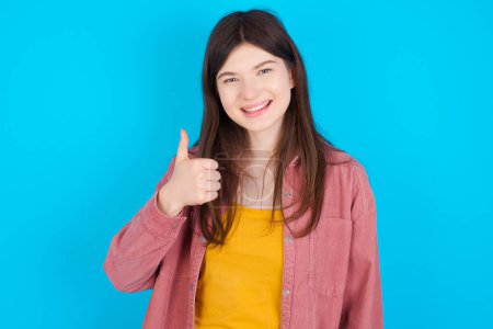 Photo for Young caucasian girl wearing pink shirt isolated over blue background giving thumb up gesture, good Job! Positive human emotion facial expression body language. - Royalty Free Image