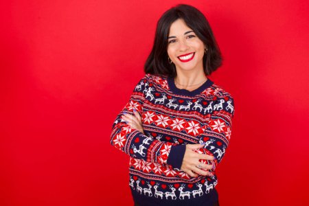 Téléchargez les photos : Brune caucasienne portant chandail de Noël sur fond rouge visage heureux souriant avec les bras croisés regardant la caméra. Personne positive. - en image libre de droit