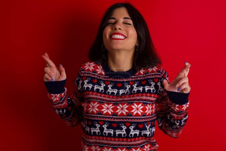 Photo for Brunette caucasian woman wearing christmas sweater over red background gesturing finger crossed smiling with hope and eyes closed. Luck and superstitious concept. - Royalty Free Image