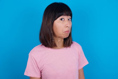 Téléchargez les photos : Jeune femme asiatique portant t-shirt sur fond bleu avec expression snob courbant les lèvres et levant les sourcils, regardant avec une expression douteuse et sceptique, suspect et doute. - en image libre de droit