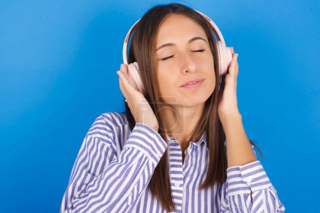Photo for Beautiful young woman with headphones on her head, listens to music, enjoying favourite song with closed eyes, holding hands on headset. - Royalty Free Image