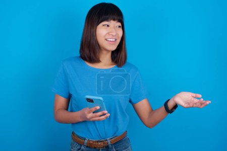 Téléchargez les photos : Heureux heureux jeune asiatique femme portant t-shirt sur fond bleu soulève la paume et détient téléphone portable utilise Internet haute vitesse pour la messagerie texte ou des appels vidéo - en image libre de droit