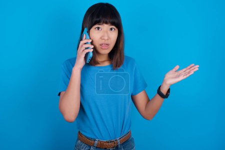 Photo for Young asian woman wearing t-shirt against blue background talking on the phone stressed with hand on face, shocked with shame and surprise face, angry and frustrated. Fear and upset for mistake. - Royalty Free Image