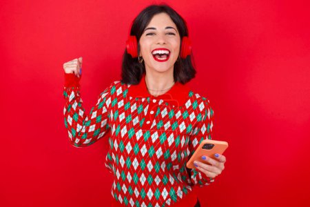 Téléchargez les photos : Femme caucasienne brune positive portant un pull de Noël sur fond rouge tient un téléphone portable moderne connecté à un casque, serre le poing de bonnes émotions, s'exclame avec joie, - en image libre de droit