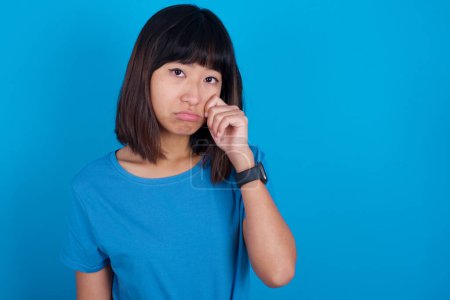 Photo for Disappointed dejected young asian woman wearing t-shirt against blue background wipes tears stands stressed with gloomy expression. Negative emotion - Royalty Free Image