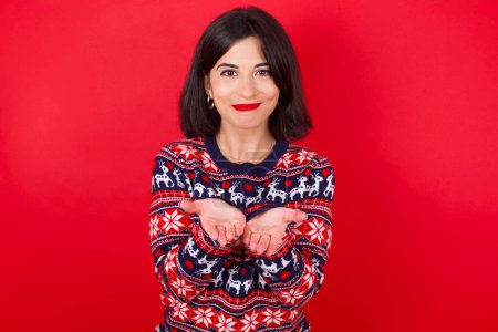 Photo for Brunette caucasian woman wearing christmas sweater over red background holding something with open palms, offering to the camera. - Royalty Free Image
