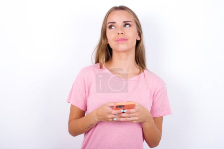 Téléchargez les photos : Heureuse jeune fille caucasienne portant T-shirt rose sur fond blanc écoutant de la musique avec des écouteurs à l'aide d'un téléphone mobile. - en image libre de droit
