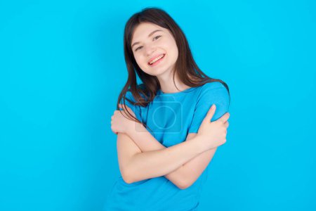 Foto de Encantadora chica caucásica joven con camiseta azul aislada sobre fondo azul abraza su propio cuerpo, se siente agradablemente cómodo plantea. Concepto de ternura y autoestima - Imagen libre de derechos
