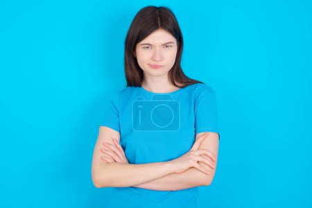 Foto de Imagen de una joven caucásica enojada con una camiseta azul aislada sobre un fondo azul cruzando brazos. Mirando a la cámara con expresión decepcionada. - Imagen libre de derechos