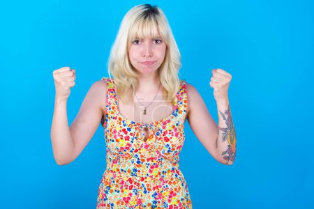 Photo for Irritated caucasian girl wearing floral dress isolated over blue background blows cheeks with anger and raises clenched fists expresses rage and aggressive emotions. Furious model - Royalty Free Image