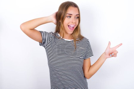 Foto de Sorprendida hermosa chica rubia con camiseta a rayas sobre fondo blanco apuntando al espacio vacío sosteniendo la mano en la cabeza - Imagen libre de derechos