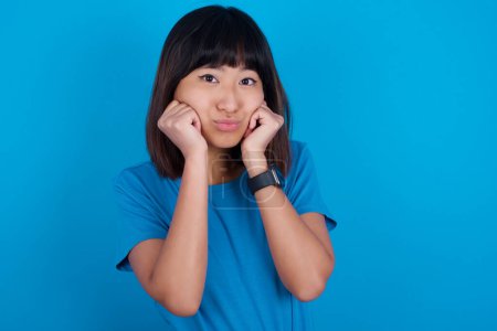 Téléchargez les photos : Jeune femme asiatique portant t-shirt sur fond bleu avec une expression surprise garde les mains sous le menton garde les lèvres pliées rend grimace drôle - en image libre de droit