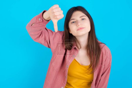 Photo for Discontent young caucasian girl wearing pink shirt isolated over blue background shows disapproval sign, keeps thumb down, expresses dislike, frowns face in discontent. Negative feelings. - Royalty Free Image