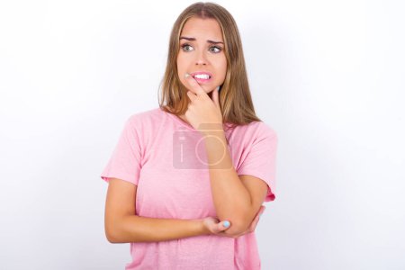 Foto de Joven chica caucásica vistiendo camiseta rosa sobre fondo blanco cubriendo la boca con las manos asustadas de algo o alguien mordiendo uñas - Imagen libre de derechos