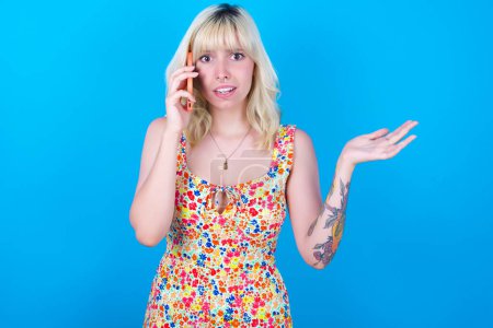 Photo for Caucasian girl wearing floral dress isolated over blue background talking on the phone stressed with hand on face, shocked with shame and surprise face, angry and frustrated. Fear and upset for mistake. - Royalty Free Image