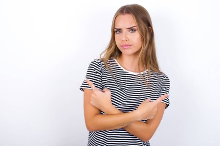 Photo for Serious beautiful blonde girl wearing striped t-shirt on white background crosses hands and points at different sides hesitates between two items. Hard decision concept - Royalty Free Image